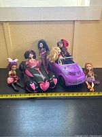 Full lot showing six Monster High dolls around two toy cars, one black with pink details, one purple convertible, measuring approximately 16 inches long.