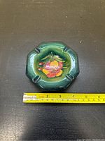 Top view of a green octagonal ashtray with a raised fruit and flower design in the center and measuring tape for scale.