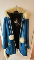 Blue parka hanging on hanger showing front with floral embroidery, fur-trimmed hood, and pom-pom drawstrings