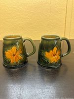 Pair of dark green glazed porcelain mugs with yellow sunflower relief on the front and rounded handles, photographed from the front on a black surface with a beige wall background.