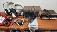 Photo showing vintage electronics on wooden table including two radios, two headphone sets, a SportStar Walkman, and battery tester with batteries.