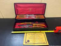 Open black fitted case showing complete set of knives, forks, and sharpening steel with amber faux stag handles inside red satin lining. Measuring tape placed in front shows length scale approximately 15 inches wide.