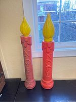 Two tall red blow-mold candle lanterns with yellow flame tops standing side by side by a window.