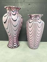 Two pink glass vases with pulled feather patterns shown front view against dark background. Taller vase on left 11" x 6", shorter vase right 9.5" x 6".