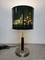 Front perspective of the table lamp showing metal base, black cylindrical stand, and lampshade with Chicago skyline at night scene.