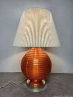Full front view of the lamp with orange Lucite ribbed spherical base, gold-toned metal pedestal base, white pleated tapered plastic shade, lamp plugged in, on grey surface against plain wall.