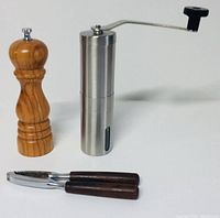 Three kitchen items displayed on a white surface including a wooden peppermill, stainless steel coffee grinder with crank, and metal nutcracker with wood handles.