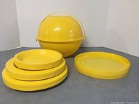 Photo showing the yellow spherical container alongside a stack of matching yellow plastic plates and an oval tray.
