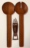 Photo shows two teak wooden salad servers, one solid spoon and one forked server, along with a fish-shaped metal bottle opener with wooden handle inset, all laid flat on a white surface.