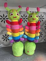 Front view of two colorful, rainbow-striped singing dancing caterpillar plush toys with green bases, pink antennae tips, and happy faces.