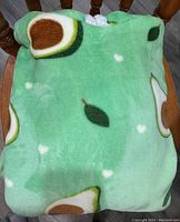 Folded avocado print blanket sweater with hood sitting on a wooden chair, showing plush green fabric with avocado pattern.