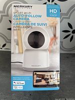 Front view of Merkury Smart Wi-Fi Auto-Follow Camera box showing the product image, brand, and key features including HD 1080P resolution and compatibility with Hey Google and Alexa.