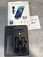 Photo of the open retail box showing two wireless lavalier microphones with foam windshields, USB-C receiver, and user manual inside black foam packaging.