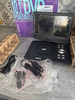 Front view of portable DVD player with screen in unfolded position, showing DVD tray and control buttons, accessories in plastic bags to front left. Original box behind.