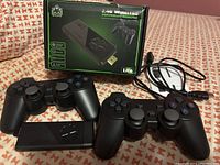 Retro game stick and two black wireless controllers laid out with USB charging cable visible. Box in background.