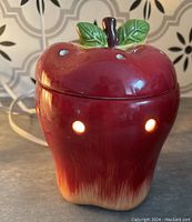 Front and side view of the red ceramic apple-shaped electric scent warmer illuminated with light glowing through vent holes.