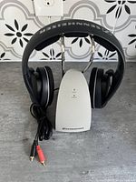 Front view of black Sennheiser wireless headphones on branded silver charger stand with audio cable visible.