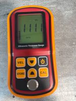 Ultrasonic thickness gauge showing a reading of 1111 on the LCD screen, sitting on a grey surface.