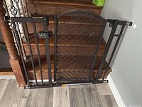 Full view of brown metal walk through baby gate installed between a wooden stair banister and a wall, showing pressure mount pads, locking latch, and decorative design on gate door panel.