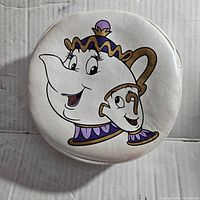 Front of white round cosmetic bag with Mrs. Potts and Chip cartoon print in purple, gold, and brown accents