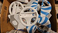 Box containing several white plastic Nintendo Wii steering wheel controllers with blue swirl designs, a white joystick attachment, and other assorted parts.