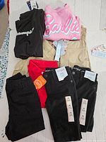 Photo of folded clothing items showing black pants, pink Barbie sweatshirt, red top, and beige pants all with tags.