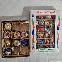 Two vintage cardboard boxes containing small assorted vintage Christmas ornaments. One box marked 'Santa Land Christmas Tree Ornaments' showing various colored ornaments inside individual compartments. Another box with dividers and paper protectors showing pink, gold, blue, and silver ornaments.