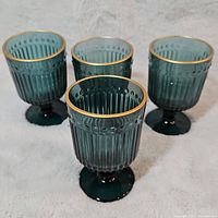 Four vintage teal glass goblets with gold rims arranged in group on a white surface, showing their shape and color.