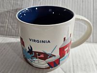 Close-up front view of the mug showing the word 'Virginia' and illustrations including a blue crab, drum, and architectural elements.