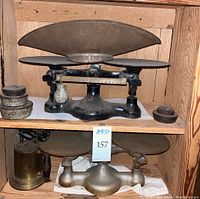 Photo showing antique candy scale with two round platforms and large curved weighing pan, cast metal weights, and brass blowtorch on wooden shelf.