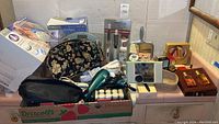 Wide photo showing entire assortment of bathroom items including hair dryers, curling iron, toiletry bags, perfumes, vintage mirror, and boxed bath mat.