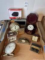 Box containing 9 assorted clocks and clock radios placed inside a cardboard box. Visible are various clock styles including travel clocks, wall clocks, and a clock radio in original packaging.
