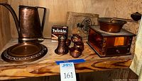 Copper items displayed on a wooden shelf: hammered watering can, round plate, coffee grinder with copper panels, small burlap bag, Twinkle copper cleaner kit, and small copper vessels