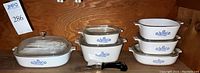 Seven white glass Corningware casserole dishes with Blue Cornflower pattern arranged on a wooden shelf, six with glass lids and one handle attachment.