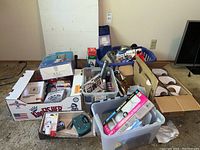 Boxes and bins of assorted household items and tools placed on garage floor with some packaging visible.