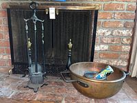 Wide shot showing entire lot including hammered copper bowl, fireplace screen, and fireplace tools on a brick fireplace hearth.