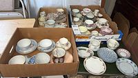 Wide view of multiple vintage teacups and saucers arranged in cardboard boxes, showing assorted styles and colors.