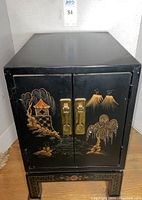 Front view of the black lacquer cabinet showing the hand-painted Chinese Chinoiserie scene on the two doors with brass hardware and decorative painted legs.