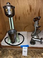 Vintage Hamilton Beach electric milkshake mixer with stainless steel mixing cup next to a Universal hand crank meat grinder with wooden handle and grill attachments, placed on cushioned surface inside a wooden cabinet.