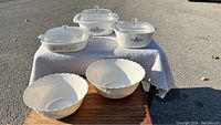 Full view of three CorningWare floral baking dishes with glass lids on a white cloth, two Arcopal white floral serving bowls placed in front on wooden surface outdoors