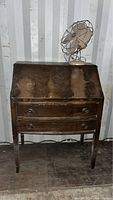 Front view of wooden drop front desk with closed writing surface and vintage electric fan placed on top