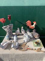 Wide photo showing multiple bird figurines of various materials arranged on a cloth, including wooden and plaster birds and decorative poppies.