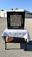 Photo of black shadow box containing custom framed Scrabble game displayed on a cloth-covered table outdoors.