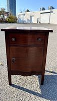 Front view of the Georgian style mahogany commode showing serpentine drawer and door, metal knobs, and keyhole, set outdoors.