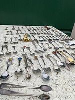 Wide view of approx 64 assorted souvenir teaspoons arranged on a white lace cloth, showing a variety of enamelled and metallic decorative tops, some with recognizable logos and symbols.