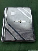 Closed view of a rectangular chrome steel cigarette case showing textured geometric pattern with diagonal herringbone stripes and a central sliding button for opening.