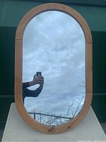 Front view of the elongated oval race track mirror with pine wood frame reflecting cloudy sky and photographer's hand holding a phone.