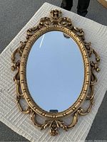 Front view of an oval mirror with an ornate gold-tone resin cast frame featuring Rococo design flourishes on a textured surface.
