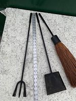 Three-piece black metal fireplace tool set (poker, shovel, broom) laid out with measuring tape showing length about 23 inches. Tools have slim handles and minimal use on broom bristles.