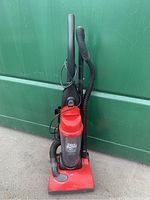 Full view of the red and black Dirt Devil upright vacuum cleaner standing upright against a green background. Shows cord wrapped around handle and attached hose.
